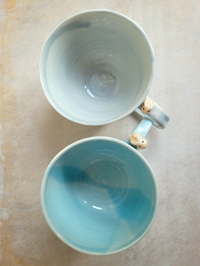 Two ceramic cups in grey and light blue with a tiny lopeared bunny on each handle flat lay