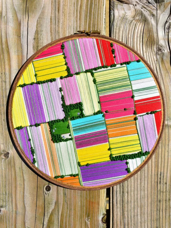 An embroidery hoop, lying on a wooden table, containing embroidered art depicting an aerial view landscape of rainbow tulip fields.
