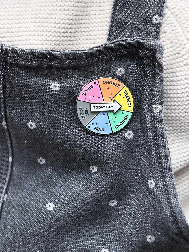 A round enamel pin that looks like a pie chart with six segments, each with positive word, and a white arrow that reads "Today I Am", is pinned to grey dungarees.