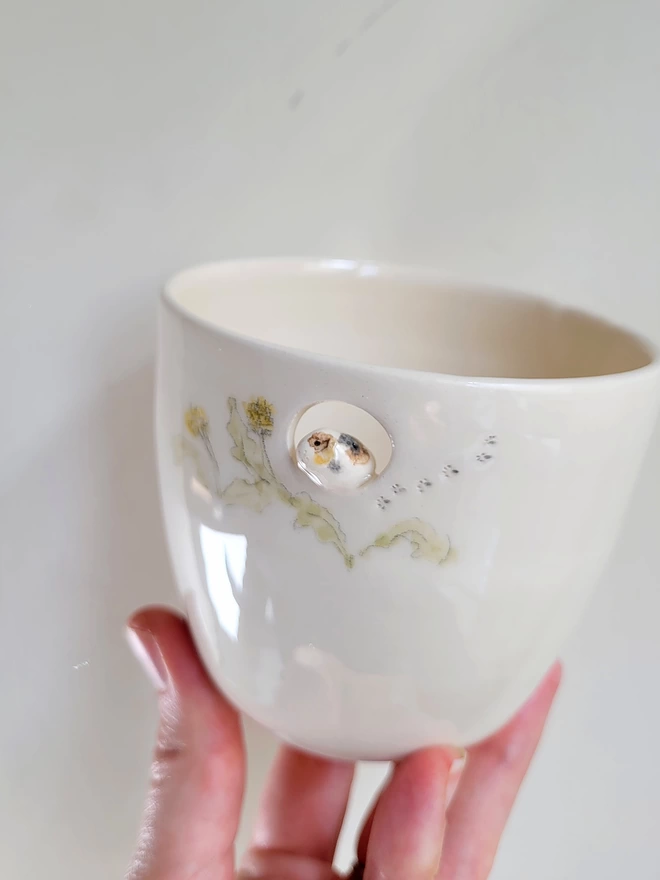 a white pottery tea cup held in hand with a tiny guineapig  sitting in a cut out hole near the rim with hand painted leaves and dandelions around it