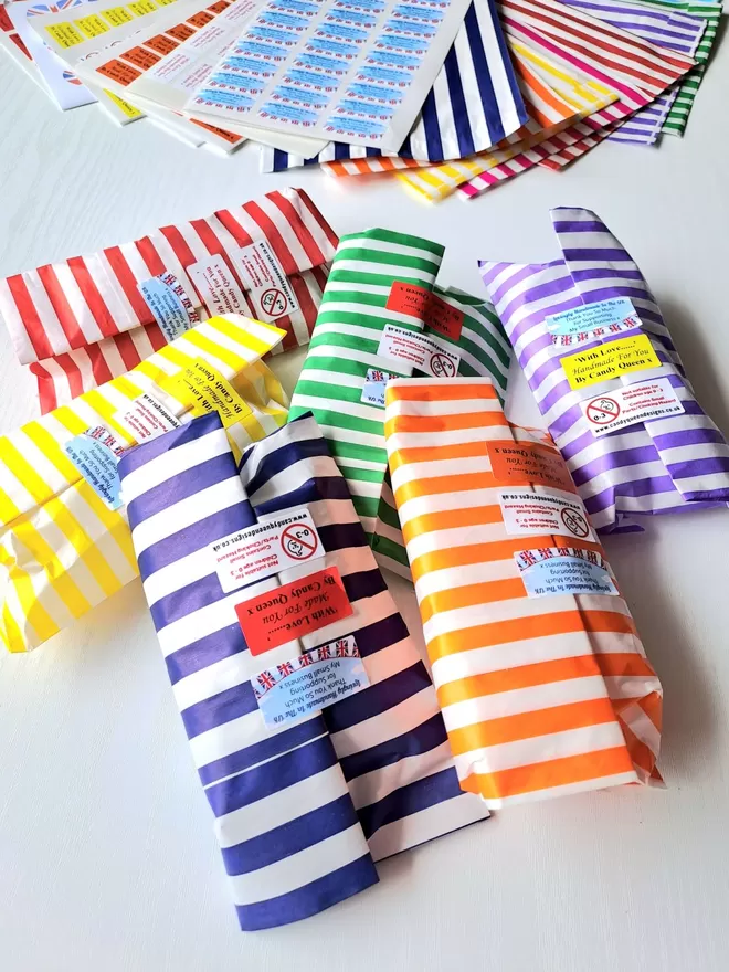 Six brightly coloured candy stripe paper bags sit in the foreground on a white worktop, the bags are red, yellow, green, blue, purple and orange. Each bag is a third full full and sealed shut with three rectangle stickers. In the background there are sticker sheets and flat bags used as packaging by Candy Queen Designs.