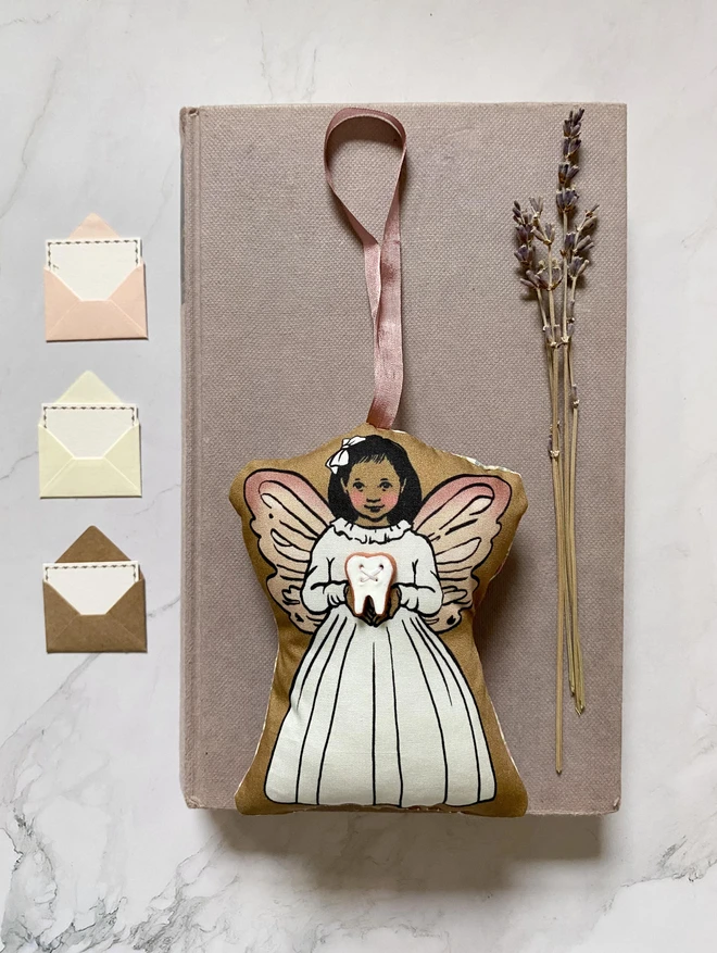 a tooth fairy plush hanging decoration laid on a vintage book with 3 envelopes next to her