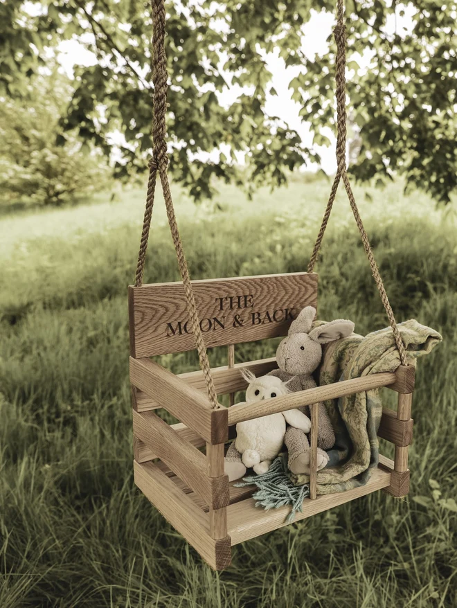 Personalised oak baby swing engraved 'The Moon & Back', styled with a soft bunny, owl and blanket, hanging from a tree in a summer meadow.