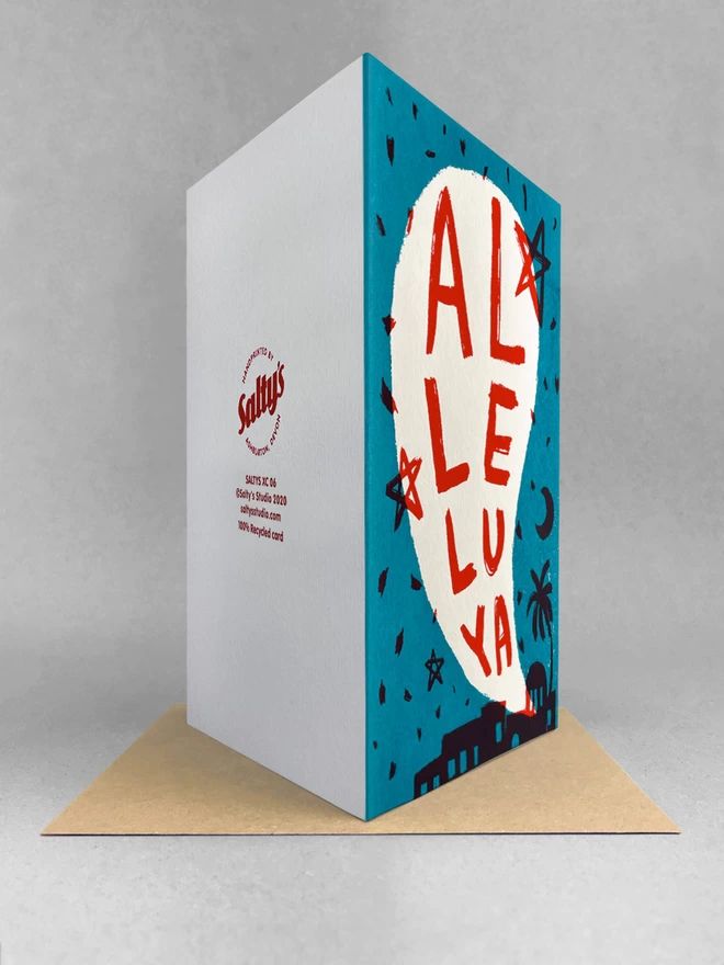 Rear view of Al-le-lu-ya handprinted Christmas card, two colour screenprint of a biblical town night scene in turquoise and red, stood on a kraft envelope in a studio setting. 