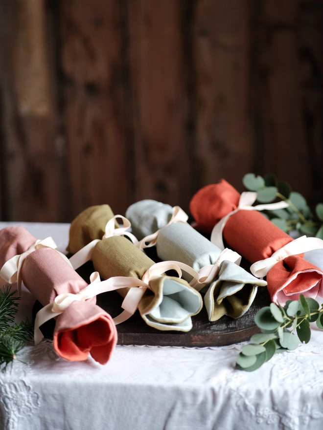 Reversible reusable linen Christmas crackers in soft pink, sage green, cream and muted festive colours, tied with natural ribbon and arranged on a family Christmas table