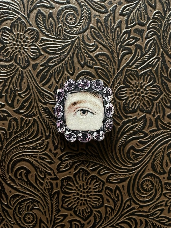 A lovers eye brooch, image of an eye painting surrounded by jewels is printed on to a wooden base