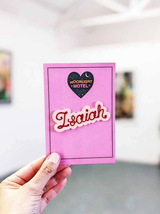 Ivory felt with Red embroidered name patch, presented on a pink backing card and held up by a womans hand, in front of a white gallery background setting