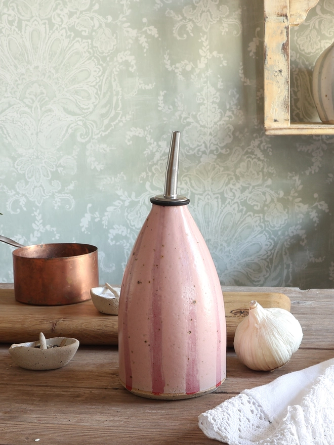 Handmade Ceramic Stoneware Oil Pourer in Pink Stripe sat on a wooden surface next to a bulb of garlic, a white vintage napkin, a brass measuring cup, a wooden chopping board and small ceramic dishes containing salt and pepper. 