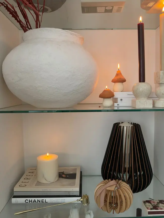 Pair Of Little Mushroom Candles (Set Of 2).  Two lit terracotta mushroom candles styled on glass shelves with a textured white vase, tall dinner candles and stacked books. The small sculptural candles add warmth and character to a styled shelf display.