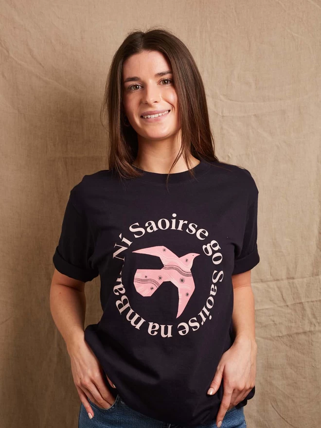 Model is shown wearing a Black & Beech navy heavyweight t-shirt with the words Ní Saoirse Go Saoirse Na mBan in pink written in a circle surrounding a pink bird