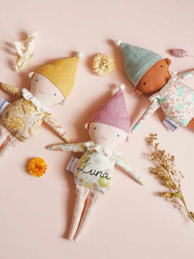 Three handmade Spring Elf Dolls are laying side by side against a soft pastel background. Each doll has a floral print body, pastel-coloured hat with a pompom, and a ruffled white collar. They feature different skin tones and gentle smiles, radiating charm and warmth.