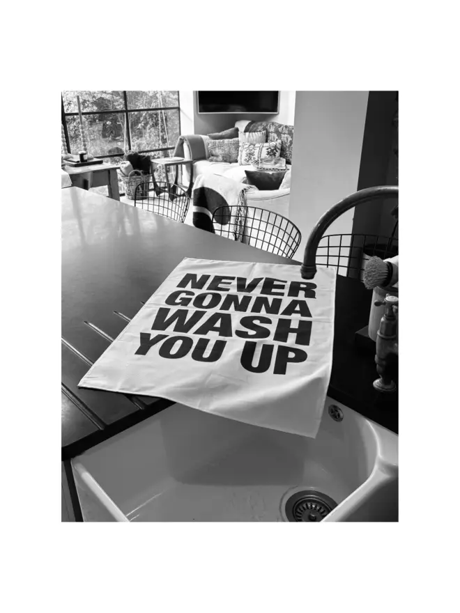 Never Gonna Wash You Up screen printed tea towel laying on kitchen counter by sink/tap with living room area in background