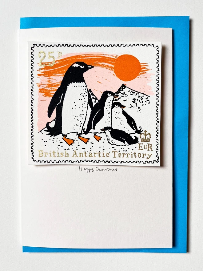 Penguin Christmas Card Sunset, a white card with a colourful penguin design in the centre on top of a blue envelope. This is placed on a white surface. 