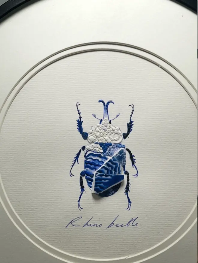 Hand-drawn, painted, mosaic clay, vintage blue Chinese Willow Tree pottery insect on white double mounted card, in a black circular frame.