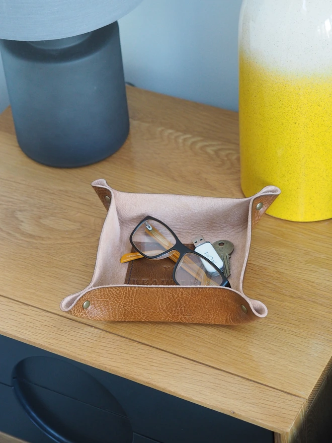 Personalised message leather coin tray. Hand dyed tan brown leather tray with a square stitched to the base which is personalised with a short but sweet message. Inside the tray is spare change, reading glasses and keys.