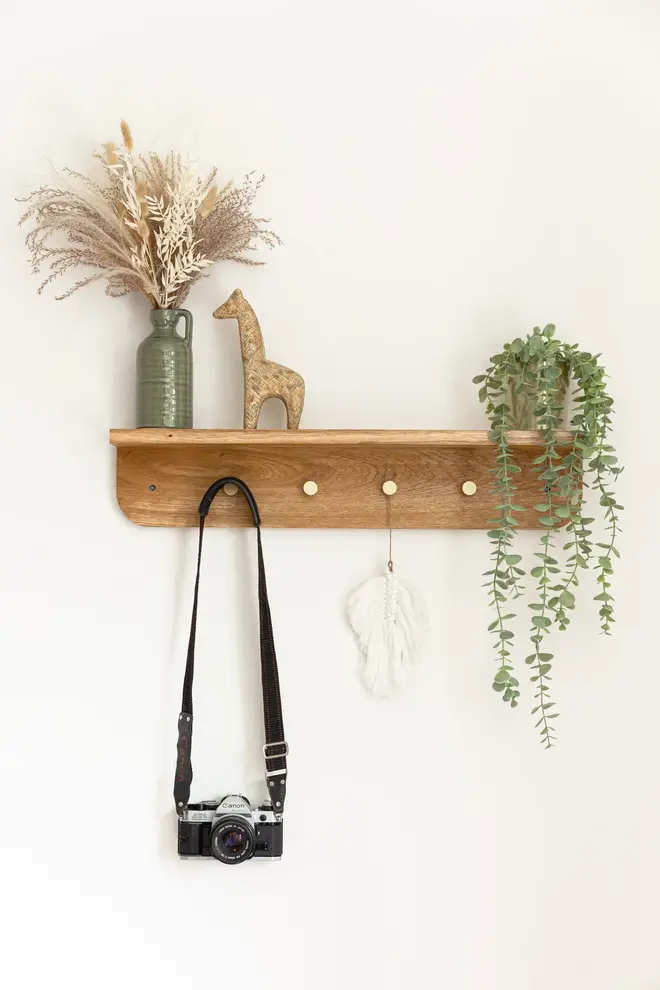 Oak Shelf Hanger, a wooden hanger hanging against a white backdrop.  It is accompanied by a vase with flowers, a camera and a plant. 
