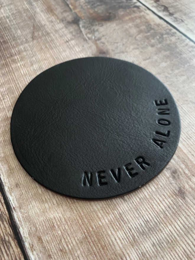 Black hand stamped leather coaster with 'never alone' stamped on it.