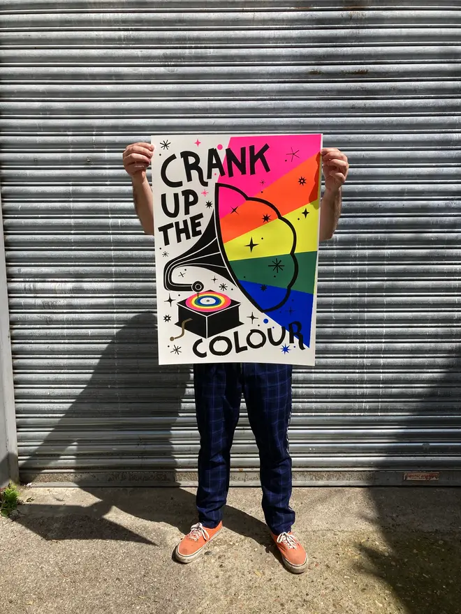 'Crank Up The Colour' Screenprint, a person holding a colourful screenprint in front of a metal shutter. The print says crank up the colour'. 
