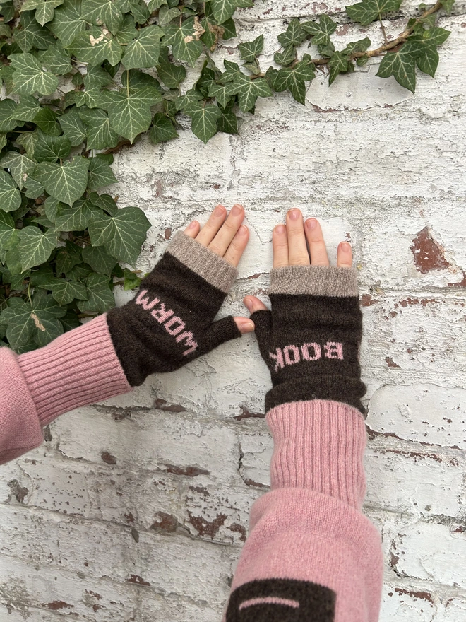 Lifestyle image of a person wearing the Cocoa & Calamine Book Worm Fingerless Mittens, with a pink jumper in front of a white brick wall with hanging leaves