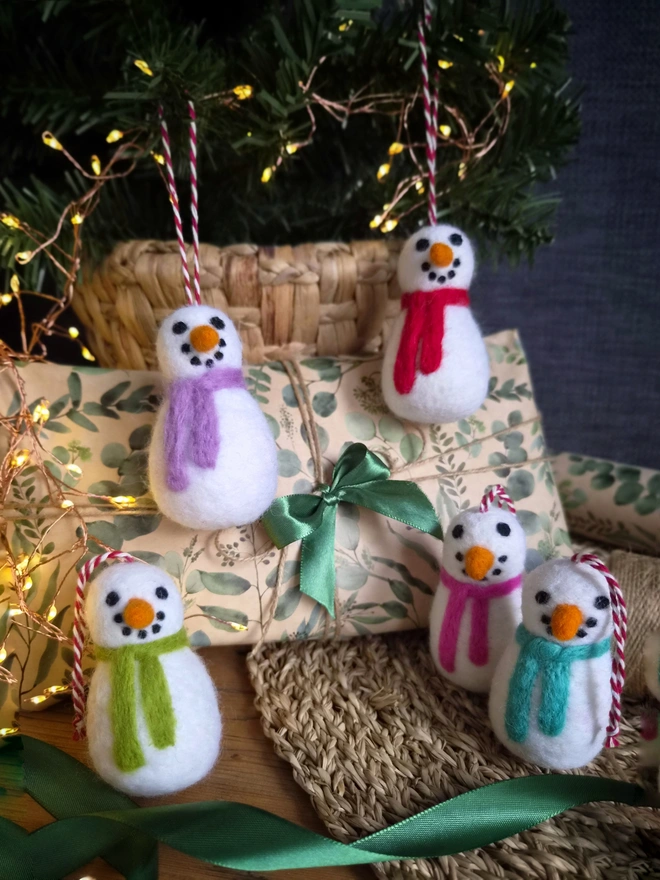 Felt snowmen tree decorations with different coloured scarves. All placed in front of a Christmas tree and present.