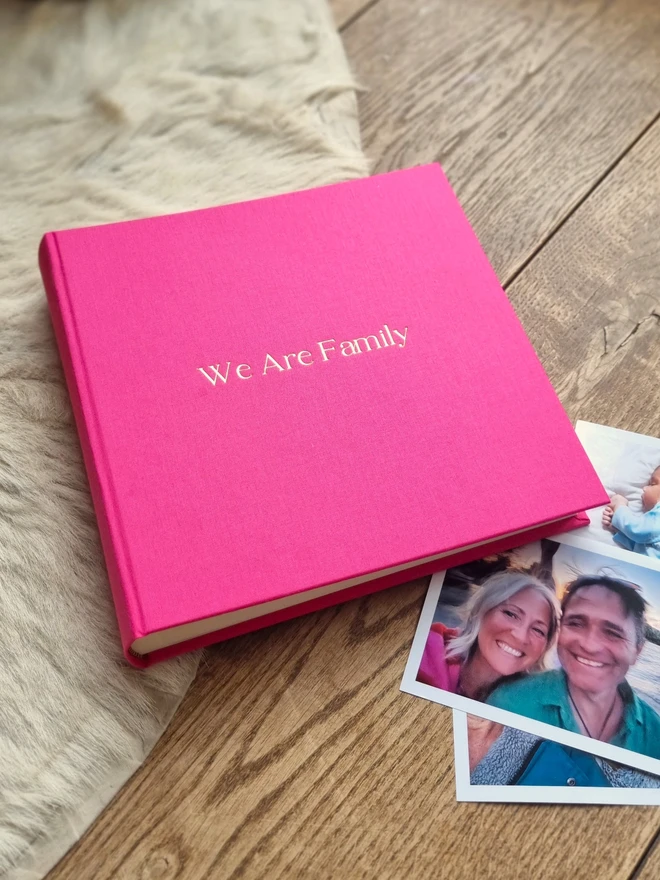 A hot pink photo album printed with 'We Are Family' in gold lettering sits on a wooden surface, next to a pile of photos.
