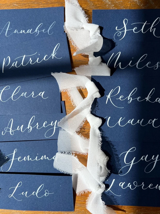 Personalised Calligraphy Wedding Navy Place Cards, navy place cards set together on a wooden table with white ribbons. 