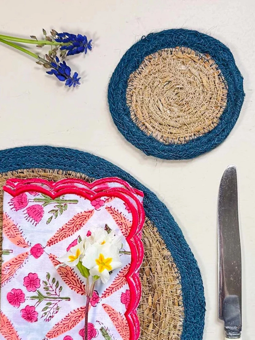 Close up of a seagrass coaster with blue rings, shown on a light surface with small white flowers. 