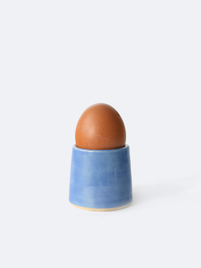 A handmade ceramic egg cup in pacific blue glaze holding a brown egg, photographed against a plain white background
