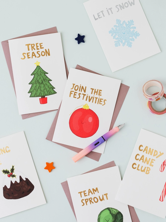 Collection of illustrated Christmas greeting cards featuring a Christmas tree, red bauble, snowflake, Christmas pudding, Brussels sprout and candy cane with gold and silver foil lettering.
