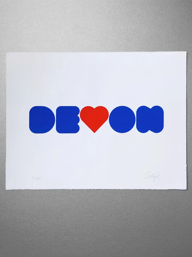 Flat lay view of a two colour screenprint, blue and red printed onto white paper. Laid on a grey background. The word Devon is written in a fat curvy font and the V depicted as a red heart