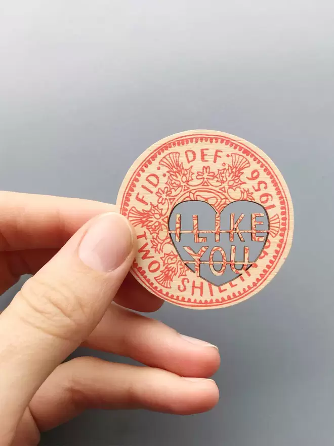 A hand holds a wooden Love token coin screen printed in red ink. The cut out words read 'I like you'