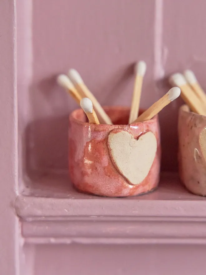 Mini ceramic match holder in pink on a pink shelf with white matches inside.