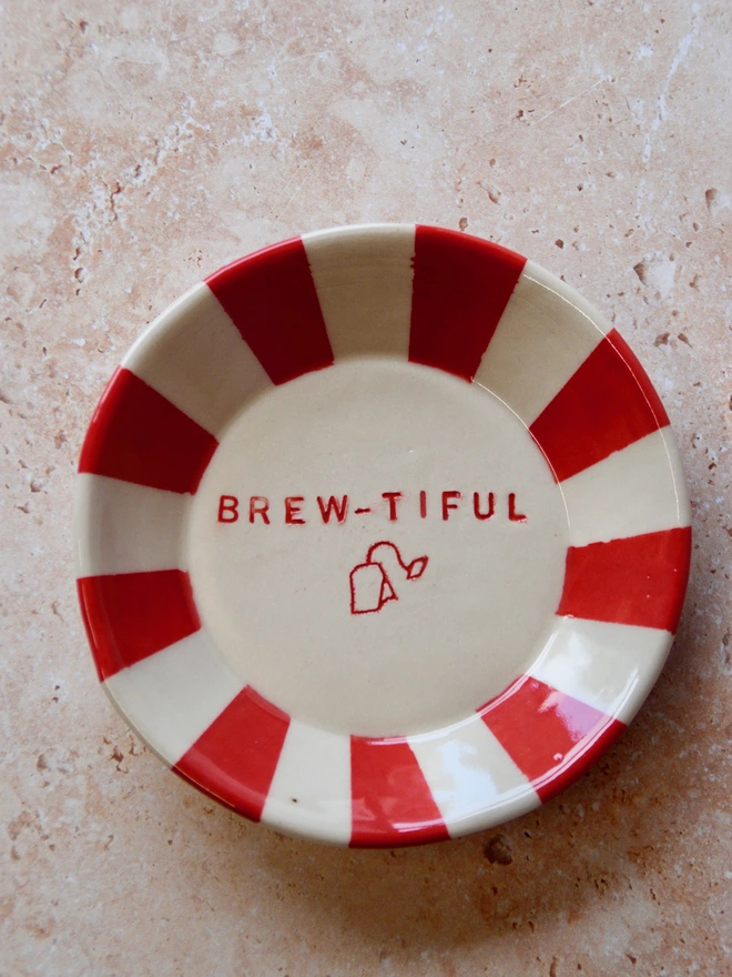 Handmade ceramic tea bag dish with red striped rim and “Brew-tiful” text with teabag illustration in the centre, on a neutral stone background.