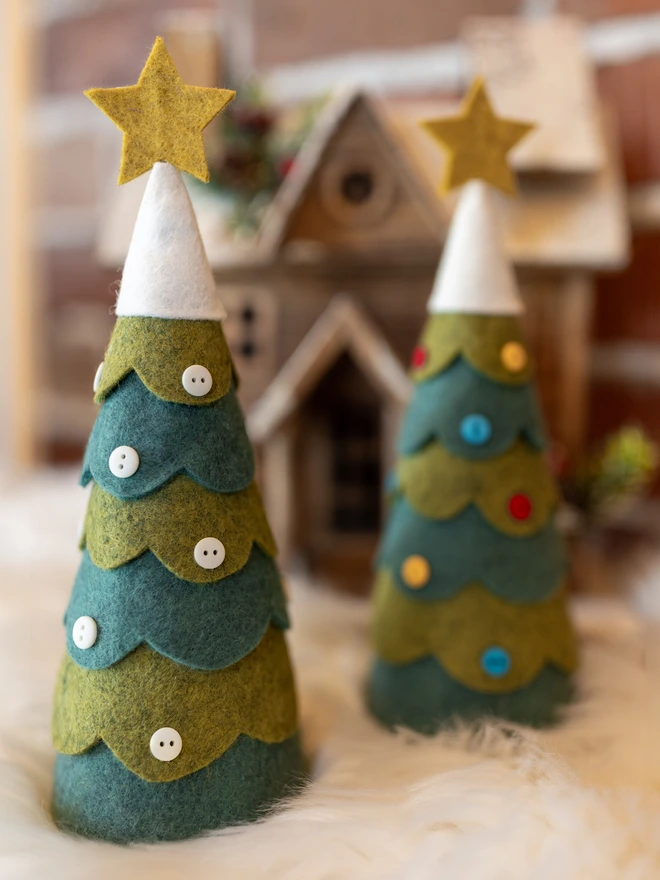 A pair of felt Christmas tree decorations. Each is decorated with buttons, one with all white, the other with red, yellow and blue. The tree has a snowy felt top adorned with a star.