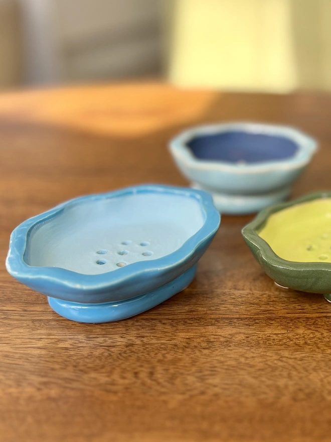 group of ceramic soap dishes on a wooden table. the dishes are colourful each in a different shade. they are oval in shape with wavy rims