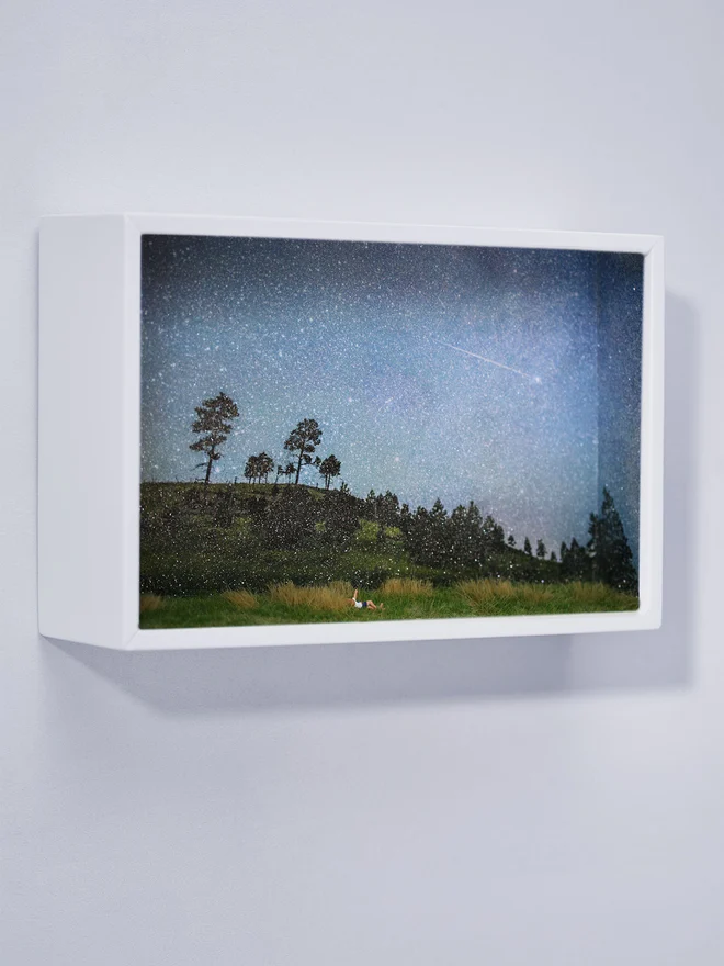 Miniature scene in an artbox showing a tiny man and woman lying in the grass watching a shooting star, against a dramatic star-filled glittery night sky.  