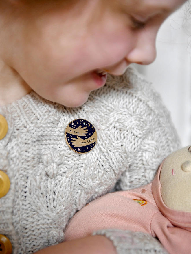 A navy blue and gold enamel pin badge with a hugging arms design and the words "A hug from Daddy" is pinned to a young child's cardigan.