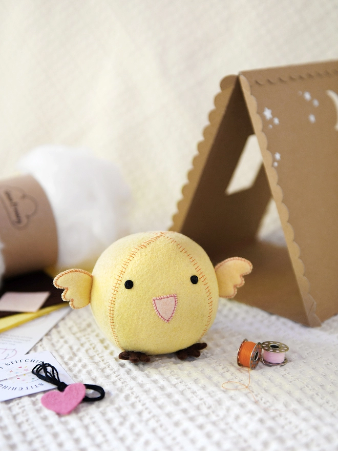 A handmade yellow felt chick toy sits beside the craft kit contents used to make it and a cardboard house.