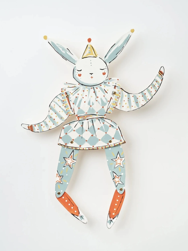 Pierrot Wall Puppet. a flat lay product shot of the pierrot wall puppet on a plain white background, showing its jointed wooden limbs painted in white, pale blue and burnt orange tones with a star motif on the tights. the hare has a closed-eye, serene expression, wearing a harlequin-style outfit with a diamond-patterned skirt and a ruffled collar, topped with a small pointed party hat. brass fasteners at the shoulders, hips and knees allow the limbs to move, giving the puppet a playful, articulated character.