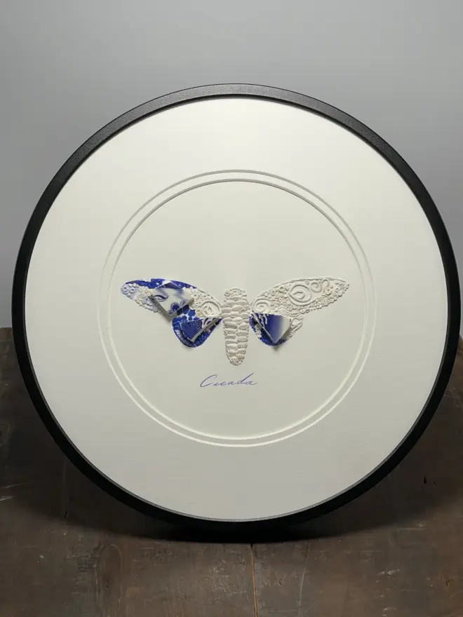 Hand-drawn, painted, mosaic clay, blue vintage Willow Tree pottery insect on white double mounted card, in a black circular frame.