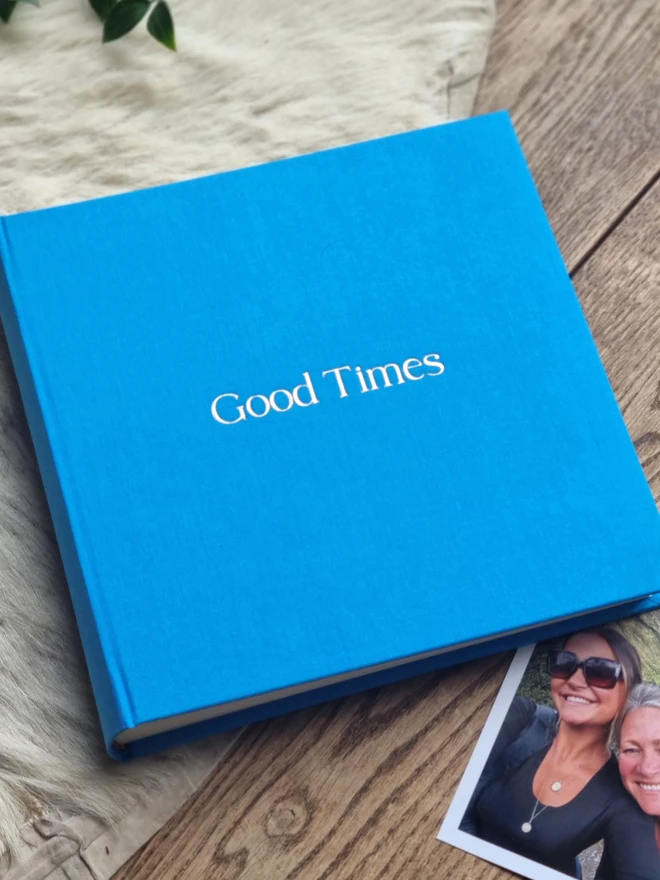 A sky blue photo album printed with the words 'Good Times' rests on a wooden floor and a rug, next to family photos.