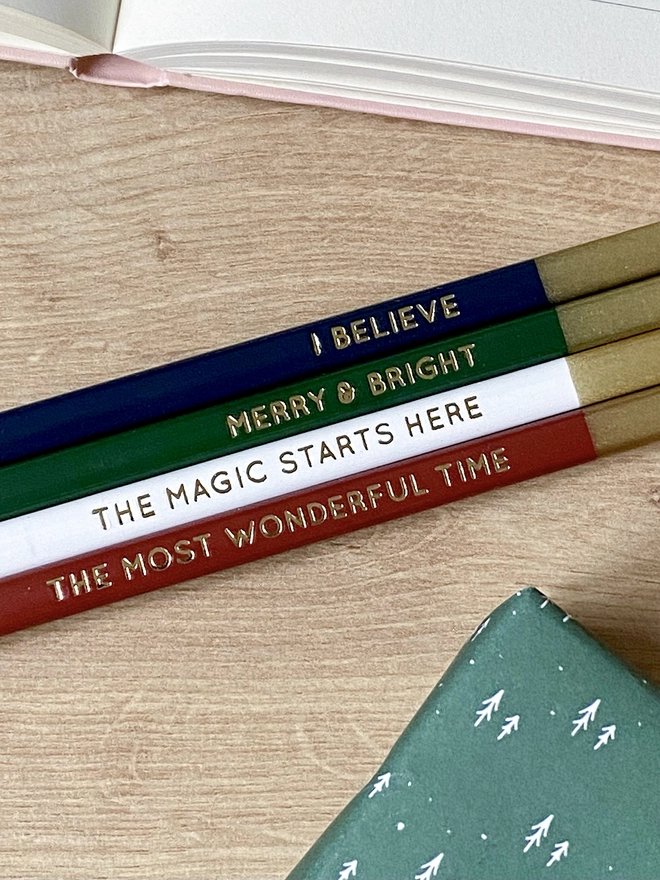 Four festive coloured pencils with gold writing along each one lay on a wooden desk beside various stationery items.
