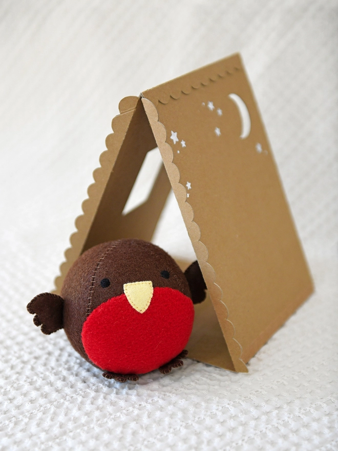 A round robin toy stands on an ivory fabric surface in front of a handmade cardboard house.