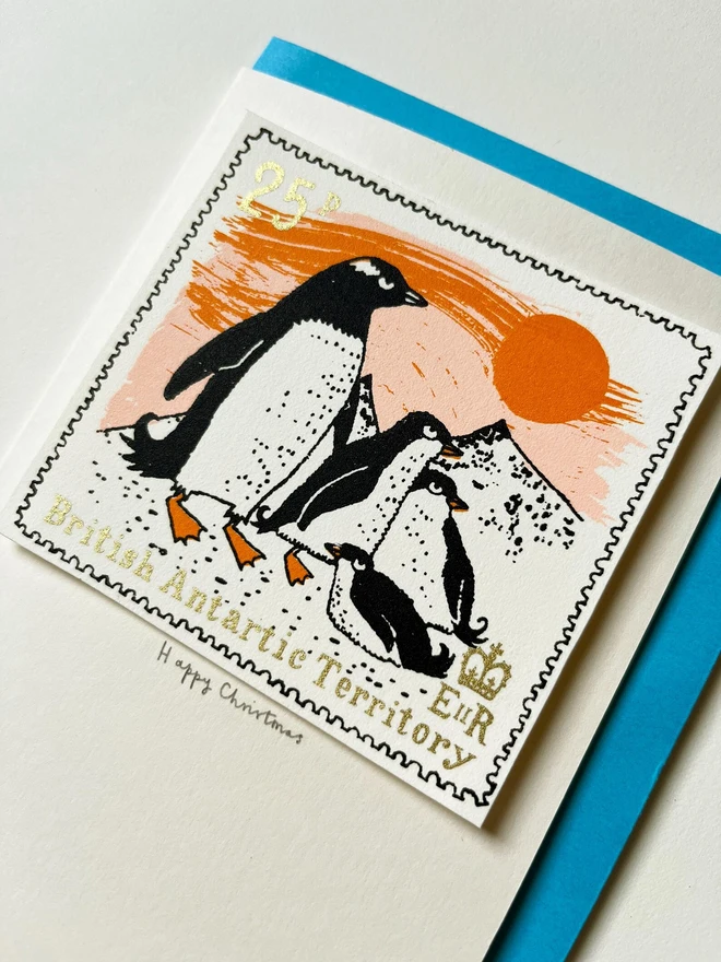 Penguin Christmas Card Sunset, a white card with a colourful penguin design in the centre on top of a blue envelope. This is placed on a white surface. 