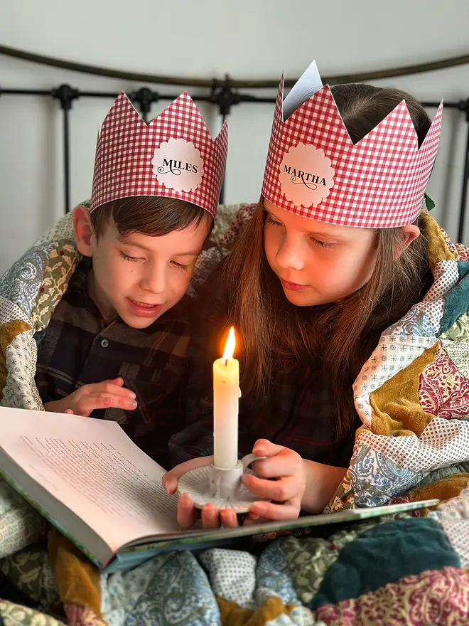 Two children wearing red gingham personalised Christmas crowns are wrapped in a cosy patchwork quilt reading a book together by candlelight.