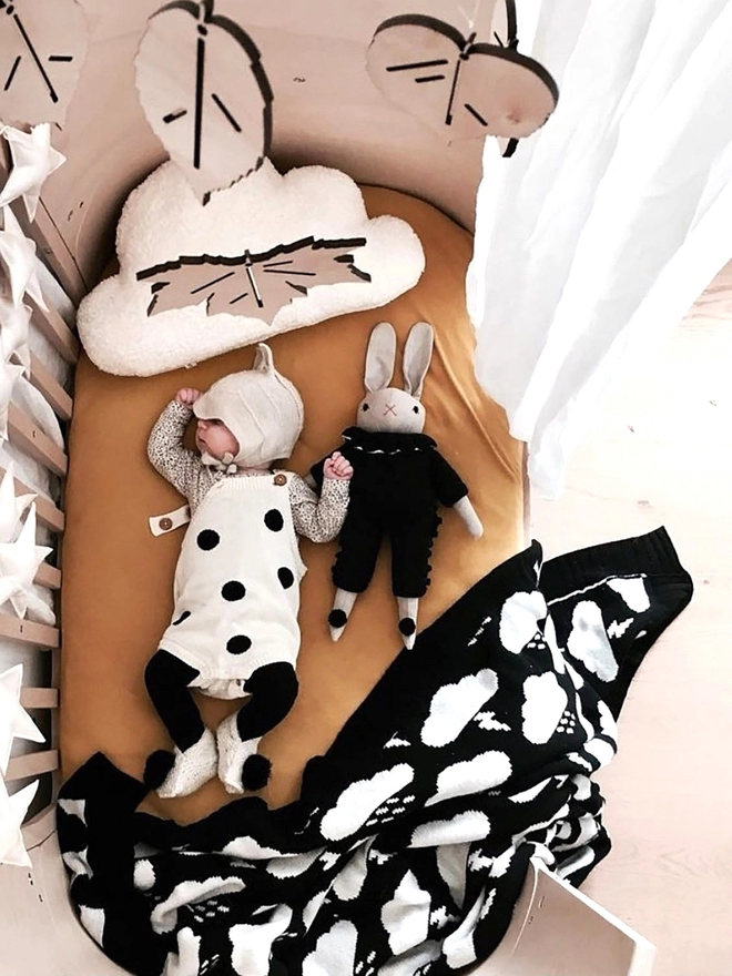 A view of a warm and neutral birch ply rounded crib with a baby asleep inside it. At the end of the crib is a black and white storm cloud blanket, next to the baby is a rabbit soft toy in a black romper. A birch plywood mobile of wooden leaves hangs above the crib.