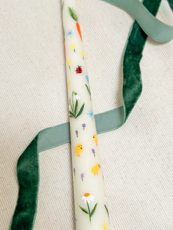 Spring Fun Candle, a white spring candle lying on a plain surface accompanied by a green ribbon. It's floral design can be seen wrapping around it. 