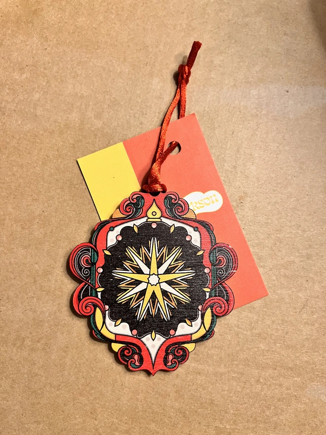 Multi coloured wooden tree decoration with a star pattern at the centre. It has a red ribbon and is lying on a brown card background with a business card with Rebecca Strickson on it. 