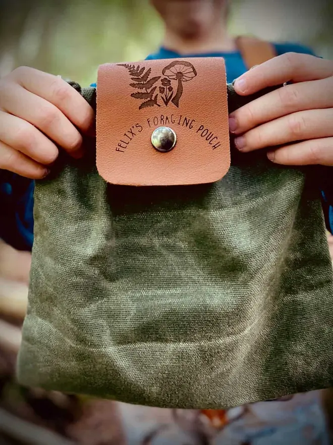 Personalised Foraging Drawstring Canvas Pouch, a child holding a green canvas pouch with a leather flap that reads "Felix's Foraging Pouch" and features drawings of a mushroom and fern.