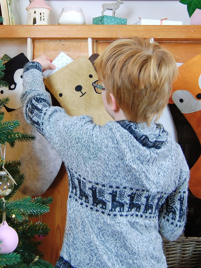 A young boy wearing a knitted cardigan is standing in front of several animal stockings hanging on a wooden mantlepiece.